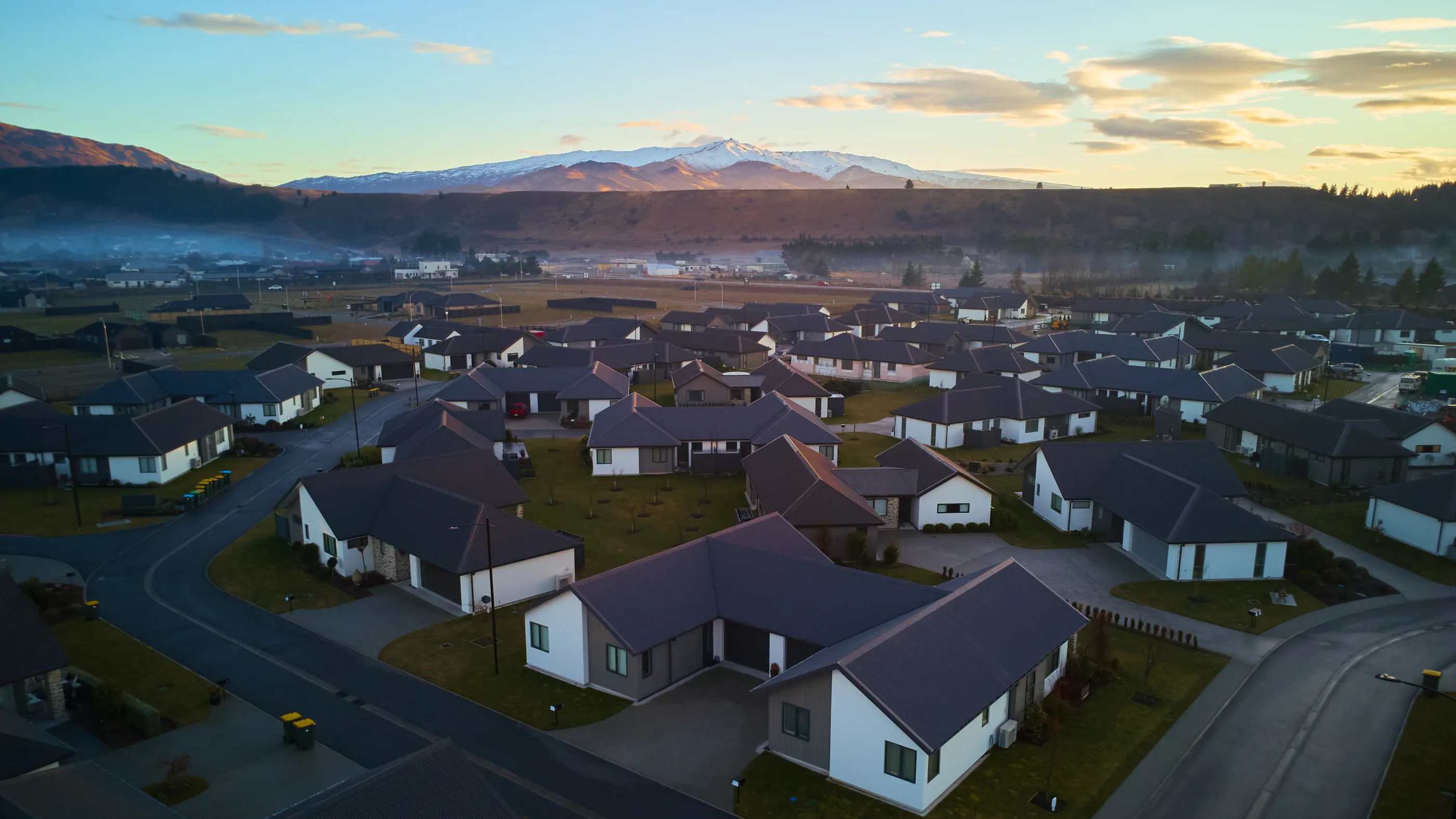 Single-storey homes in the foreground, a township and low mist beyond them, and a snow-capped range along the horizon at dusk.
