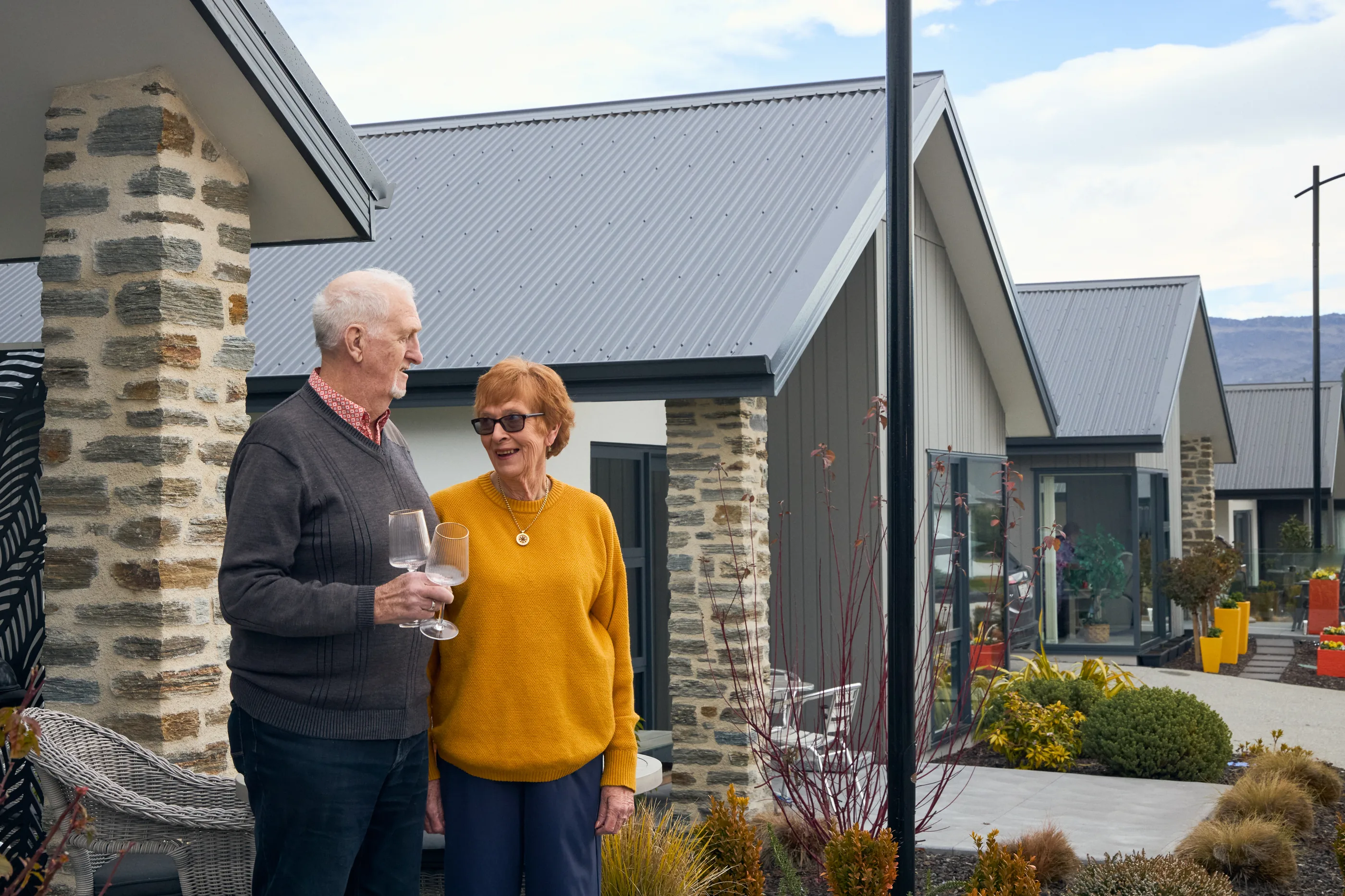 A couple standing together outside their front door with a glass each, schist piers beside them and village rooflines running back toward the range.
