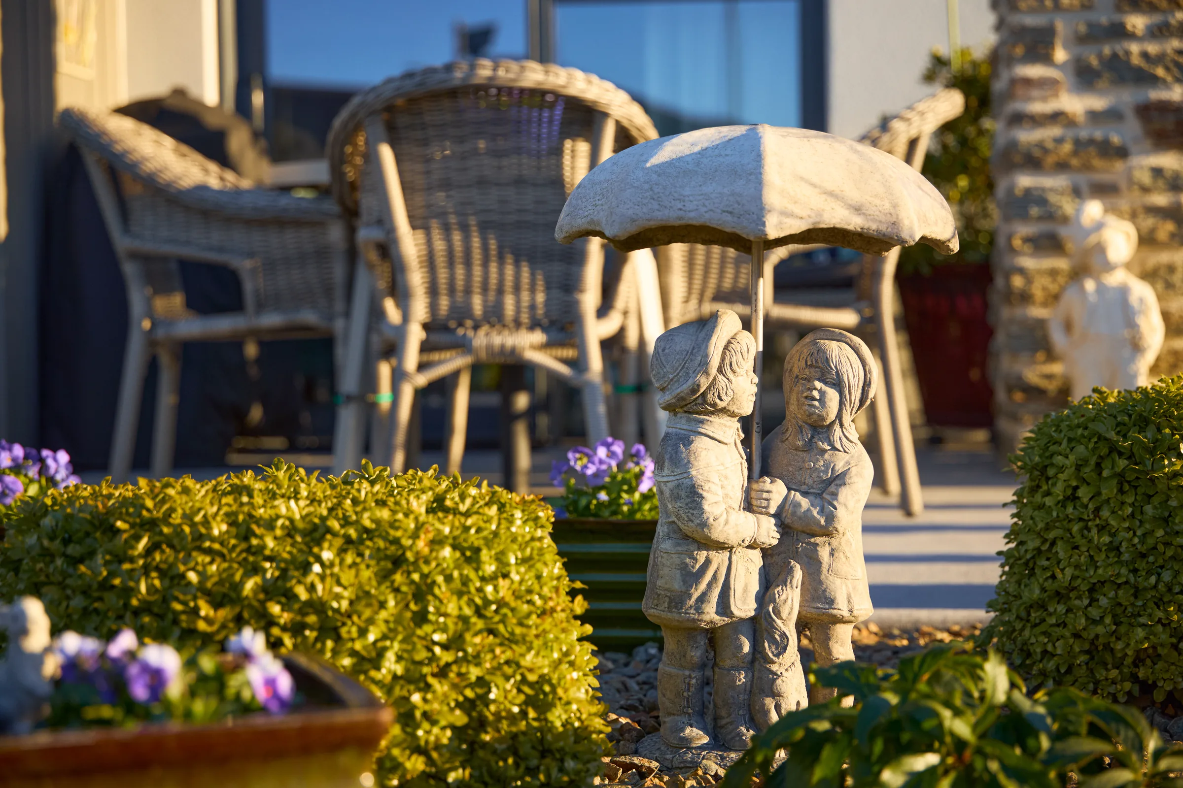 Two stone garden figures on a gravel bed, wicker chairs behind them.