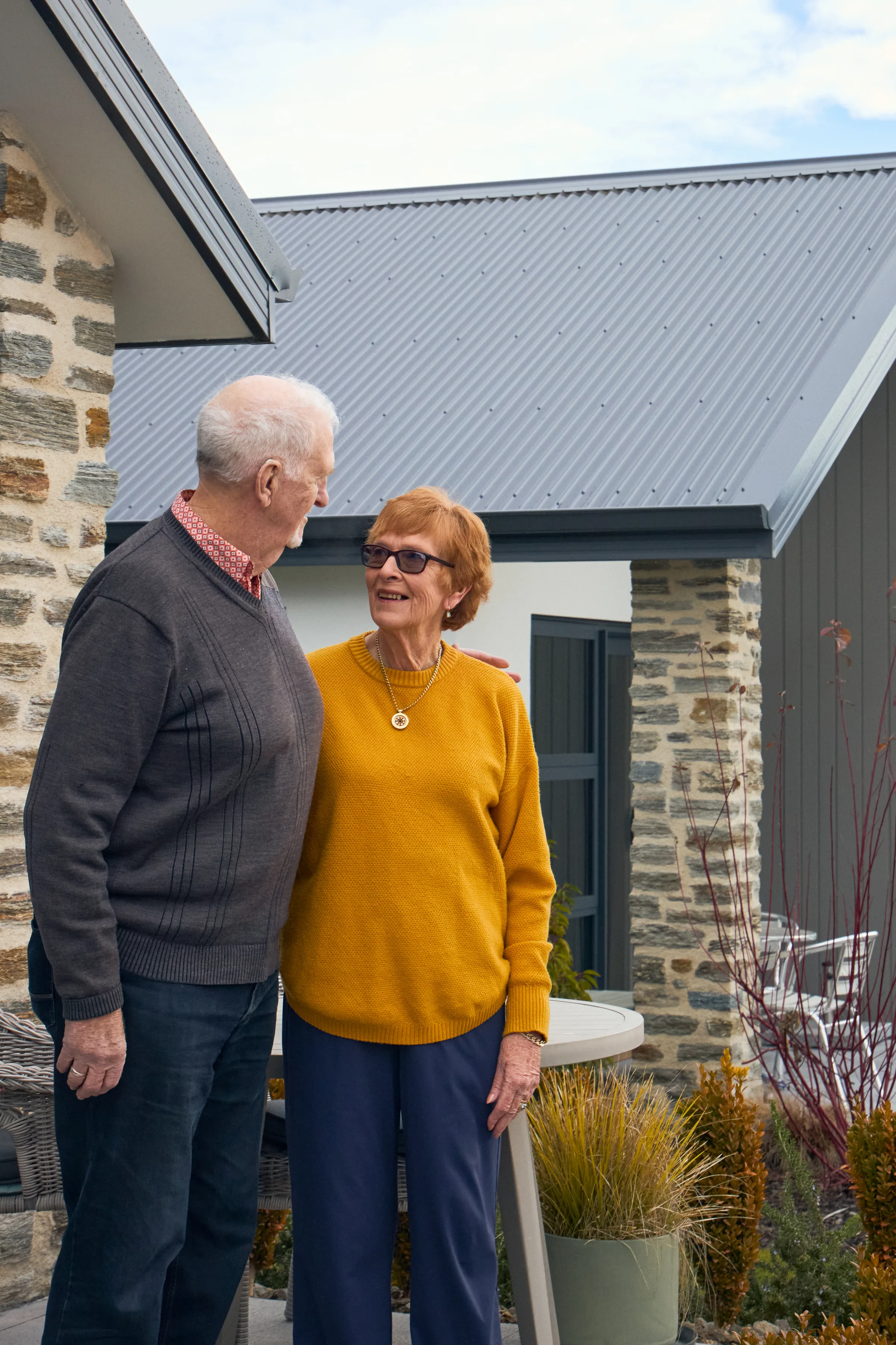 A couple laughing together on the doorstep of their home, a schist pier beside them.