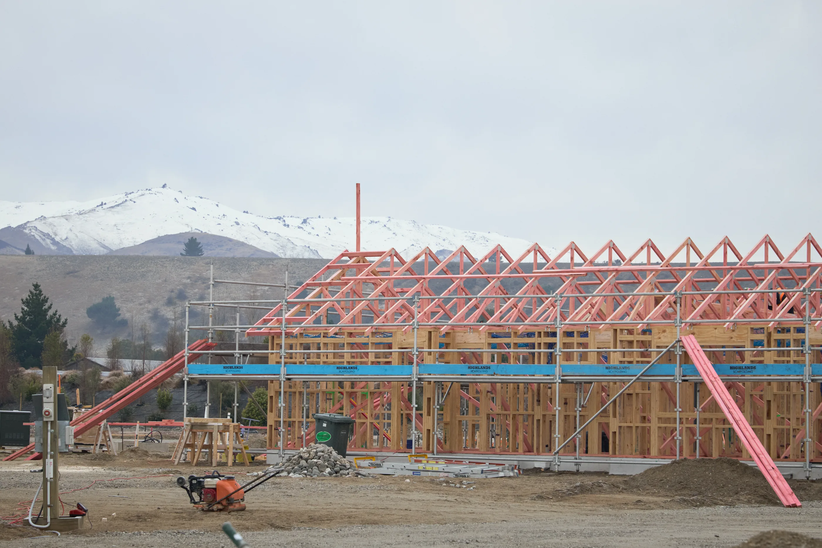 Exposed roof trusses along the length of a building under construction, against a snow-capped range.