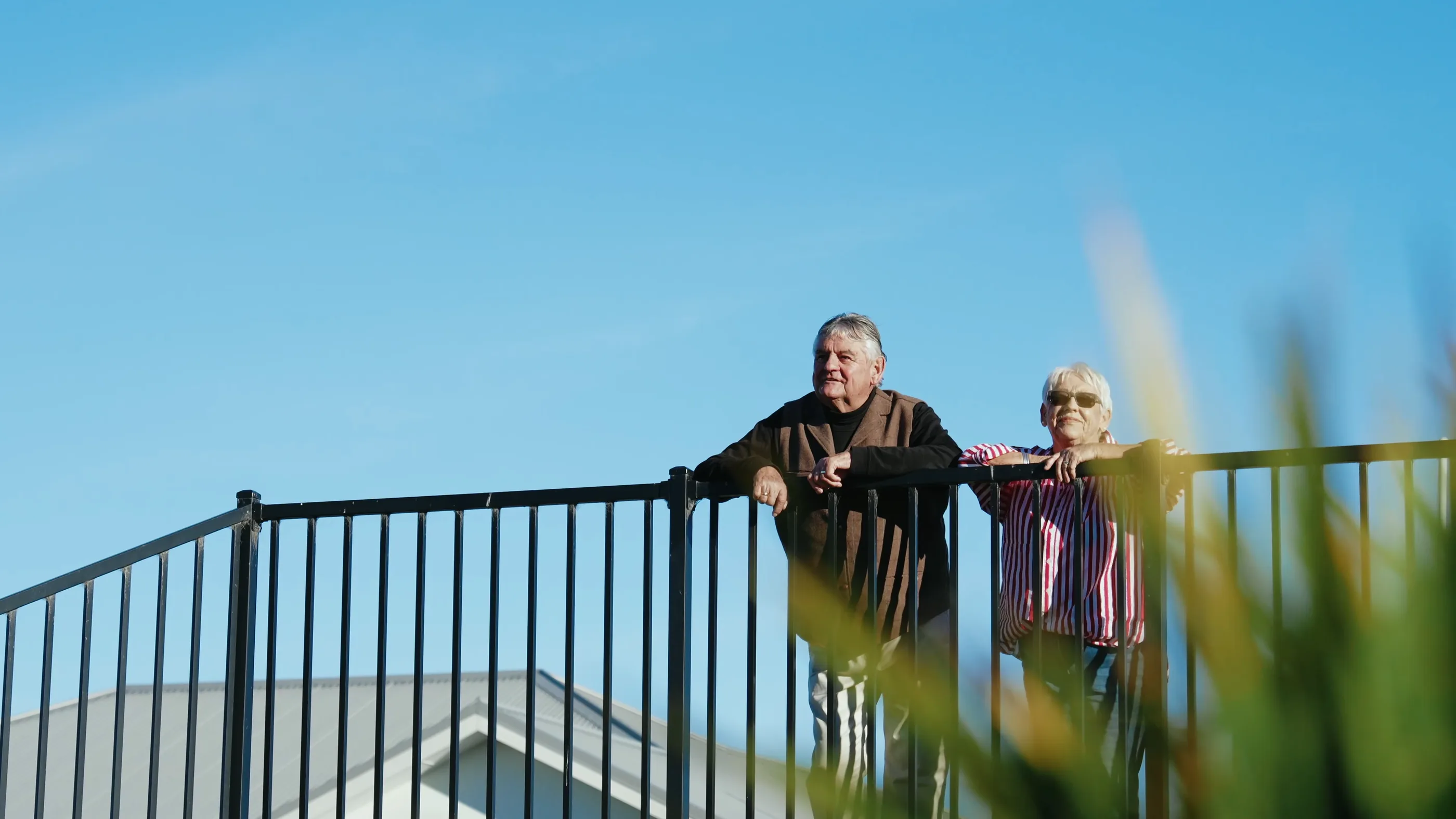 A man and a woman lean on a railing together, looking out past the garden, both smiling in the afternoon sun.