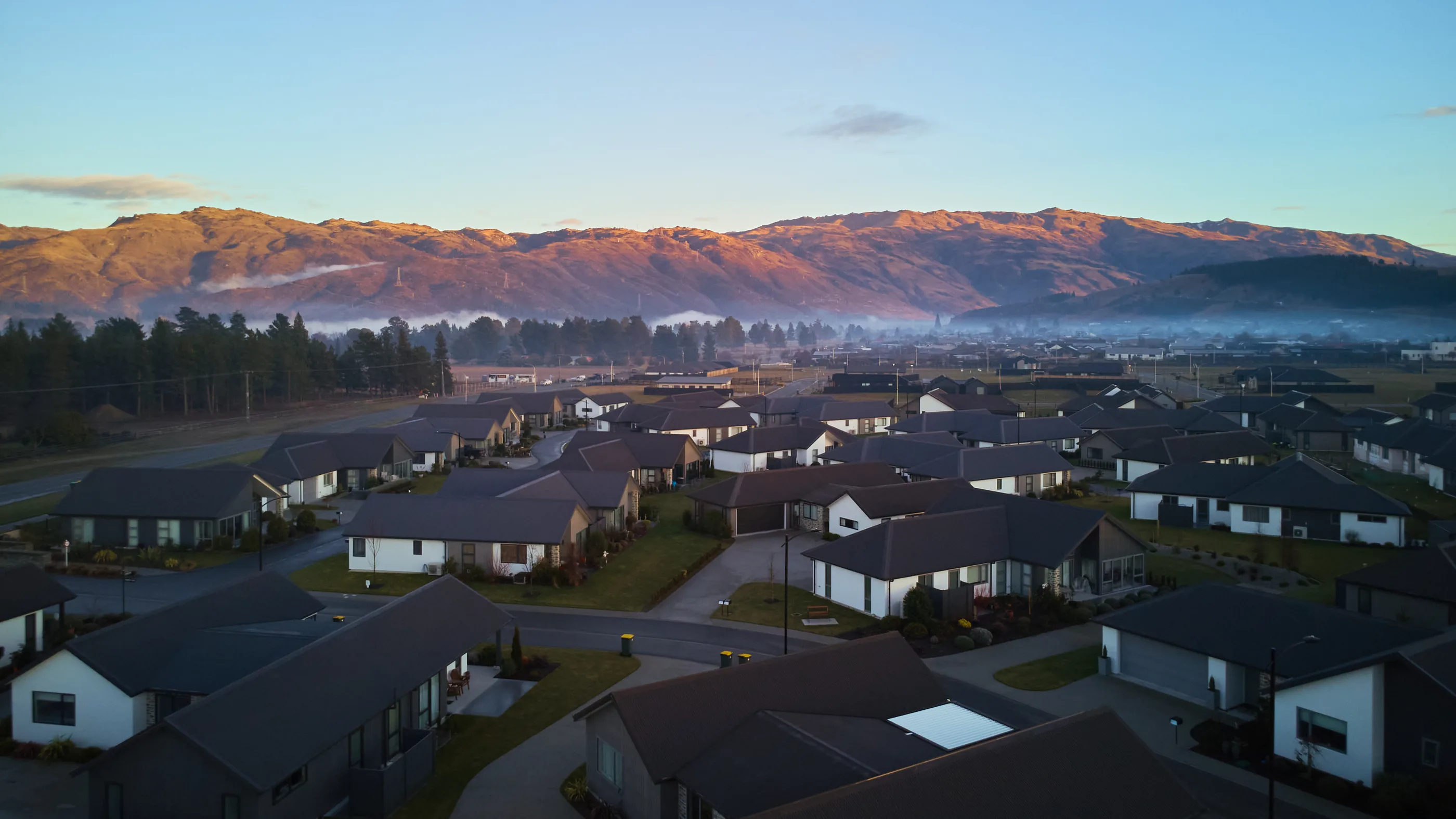 An aerial view of village homes at dawn, with ground fog lying across the valley behind them and first light on the range above.