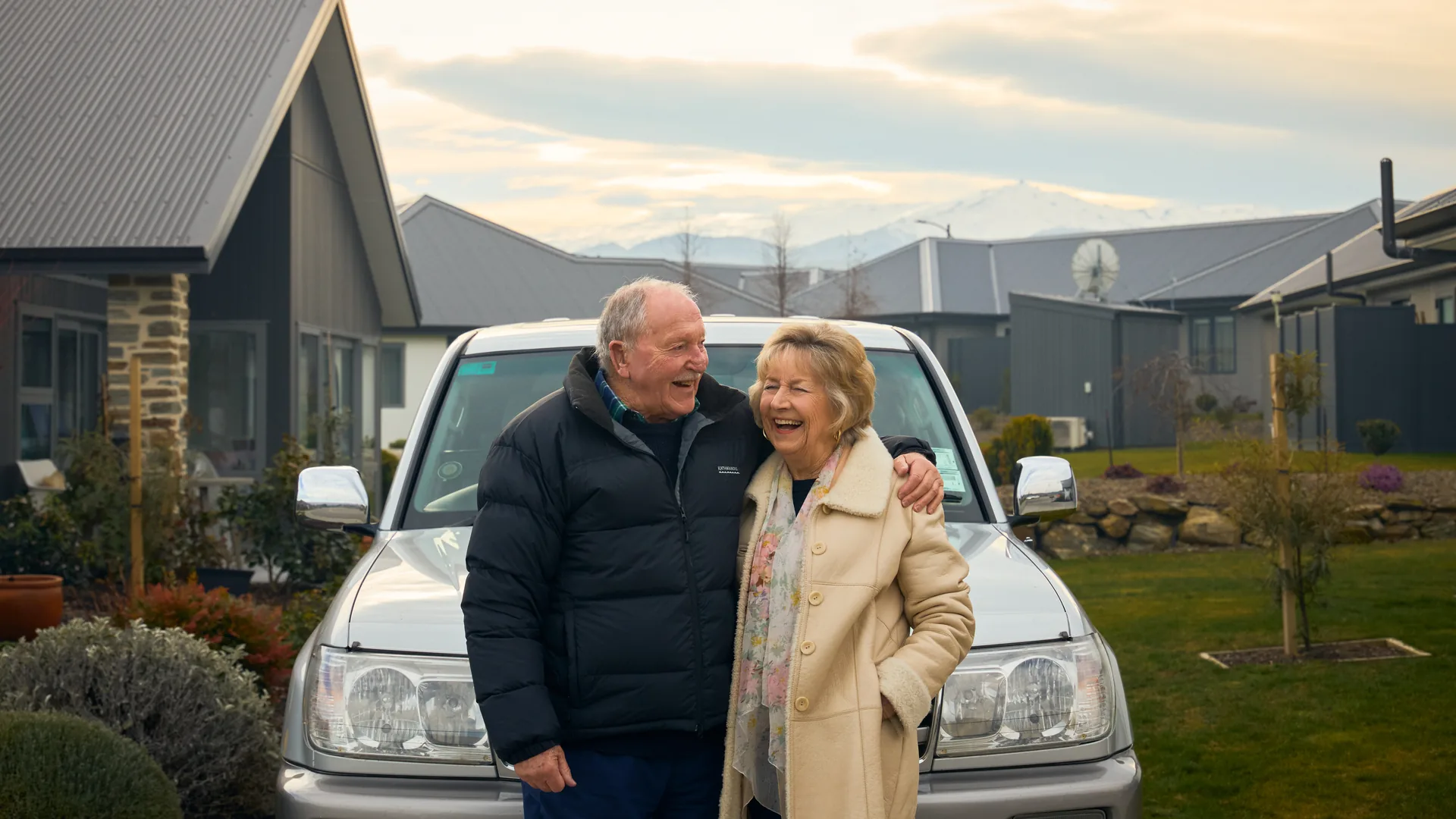 A couple laughing outside a villa at Aurum on Clutha in Clyde, with the snow-covered range behind.