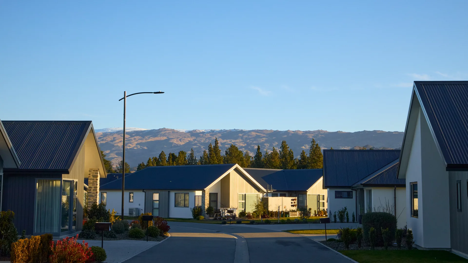 Completed villas either side of a street at Aurum on Clutha in Clyde, with the Central Otago range behind in low evening light.