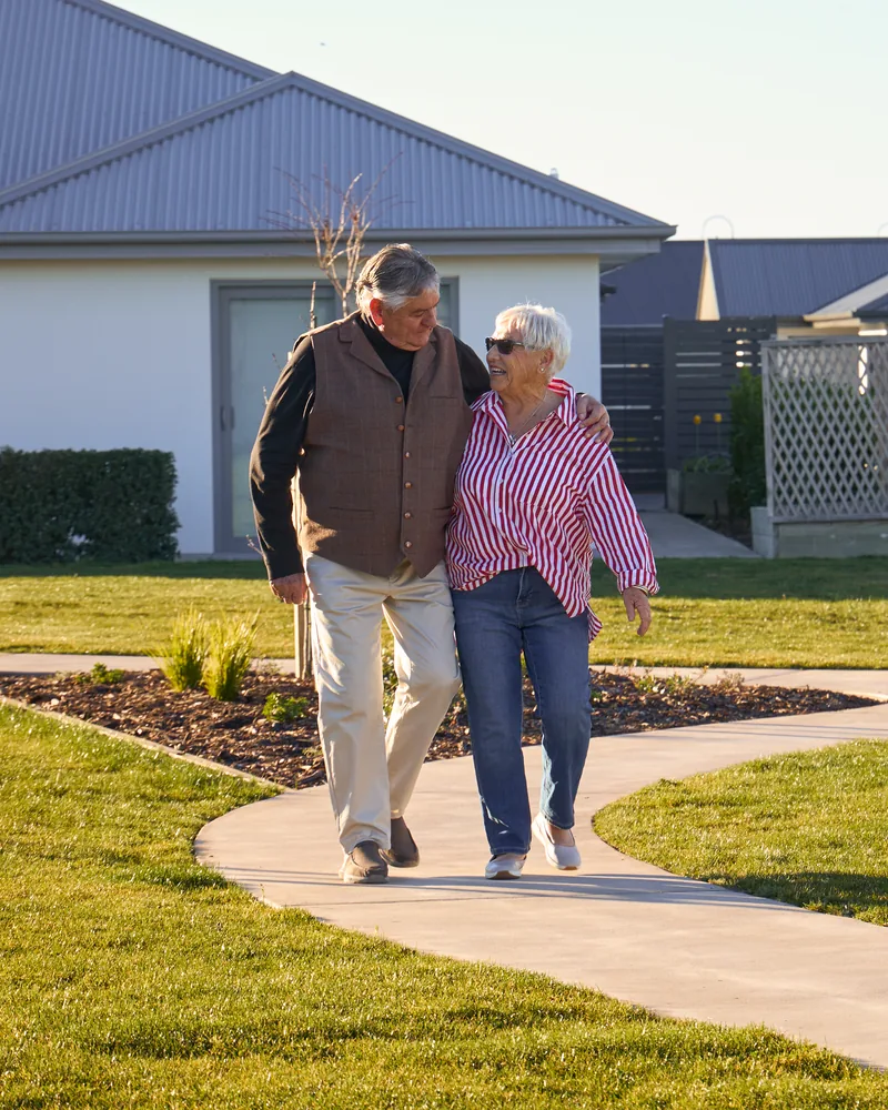 A couple walking arm in arm along a path between villas, in low evening light.