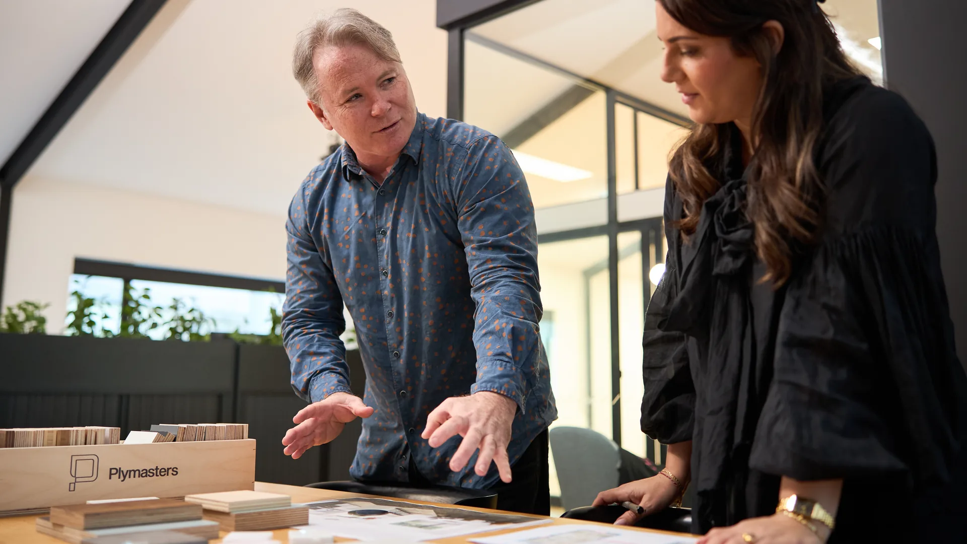 Two people talking over material samples for a village, plywood swatches and drawings laid out on the table between them.
